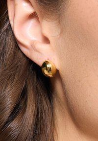 Złoty kolczyk wkręcany wyeksponowany na uchu, o gładkim, zaokrąglonym kształcie i lustrzanej powierzchni, na tle ciemnych, brązowych włosów.