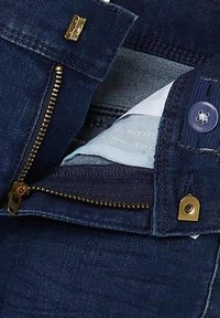 Los jeans de mezclilla azul oscuro cuentan con una cremallera dorada, cierre de botón y un forro de tela azul claro visible en el interior.