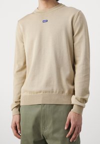 Beige crewneck-paita, jossa on ribbikäsivarret ja helma, ja rinnassa pieni sininen "HUGO"-logo. Valmistettu sileästä, hienoista neulemateriaalista.