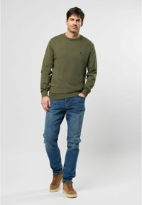 Pulover tricotat de culoare verde măsliniu, cu guler rotund, asortat cu blugi din denim albastru. Modelul poartă pantofi înalți de culoare bej. Puloverul are o textură fină de tip ribbed.