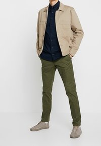 Chaqueta ligera beige con dos bolsillos frontales sobre una camisa azul marino, combinada con pantalones verde oliva y zapatillas beige claro.