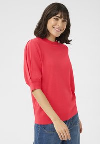 Pull en tricot rouge avec un col montant et des manches courtes bouffantes. Présente un ourlet et des poignets côtelés, porté avec un jean en denim bleu.