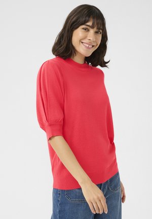 Pull en tricot rouge avec un col montant et des manches courtes bouffantes. Présente un ourlet et des poignets côtelés, porté avec un jean en denim bleu.