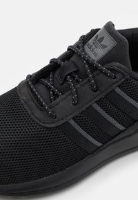 Black athletic shoe with mesh upper, suede accents, and striped design. Features dark laces and a branded tag on the tongue.