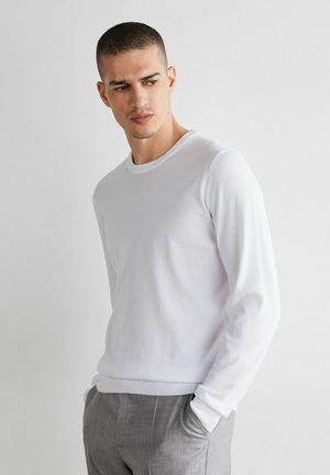 Mann mit kurzem Haar, der ein weißes Langarmhemd und graue Hosen trägt, steht mit den Händen in den Taschen vor einer schlichten Wand.