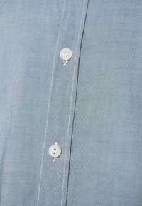 Valkoinen nappitusviimeistely ja valkoiset saumat kevyessä sinisessä puuvillapaidassa.