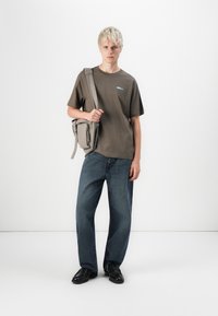 Olijfgroene t-shirt met een blauw logo, losse blauwe jeans, zwarte schoenen en een kleine grijze schoudertas met een rits.
