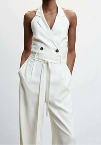 Mono blanco con cuello halter, diseño de doble botonadura, botones marrones, bolsillos laterales y cintura con cinturón para un ajuste ajustable. Textura suave.