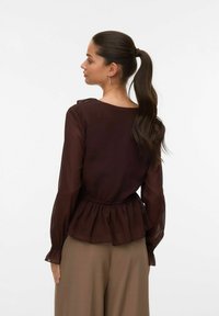 Smeđa chiffon bluzica s dugim rukavima i elastičnom gumicom u struku, s naglašenim peplum krojem i širokim izrezom. Uparena s bež hlačama.