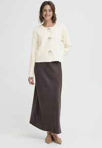 Ivory pleteni kardigan s tremi srebrnimi zavihki, v kombinaciji s dolgim, temno rjavim satenastim krilom. Ima gladke teksture in sproščen kroj.