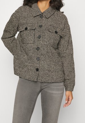 Femme portant une veste boutonnée à motif pied-de-poule noir et blanc avec des poches à rabat et un jean gris sur un fond uni.