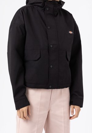 Le modèle porte une veste noire à capuche avec des boutons-pression et des poches à rabat, assortie à un pantalon rose pâle.