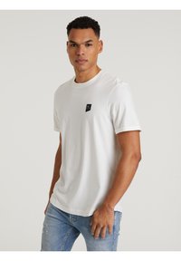 Weißes kurzärmliges Baumwoll-T-Shirt mit rundem Ausschnitt und einem kleinen schwarzen Logo-Patch auf der linken Brust. Getragen mit hellblauen, zerrissenen Jeans.