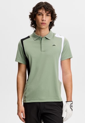 Giovane uomo che indossa una polo verde chiaro con pannelli neri e bianchi, pantaloni neri e un guanto da golf bianco sulla mano destra.