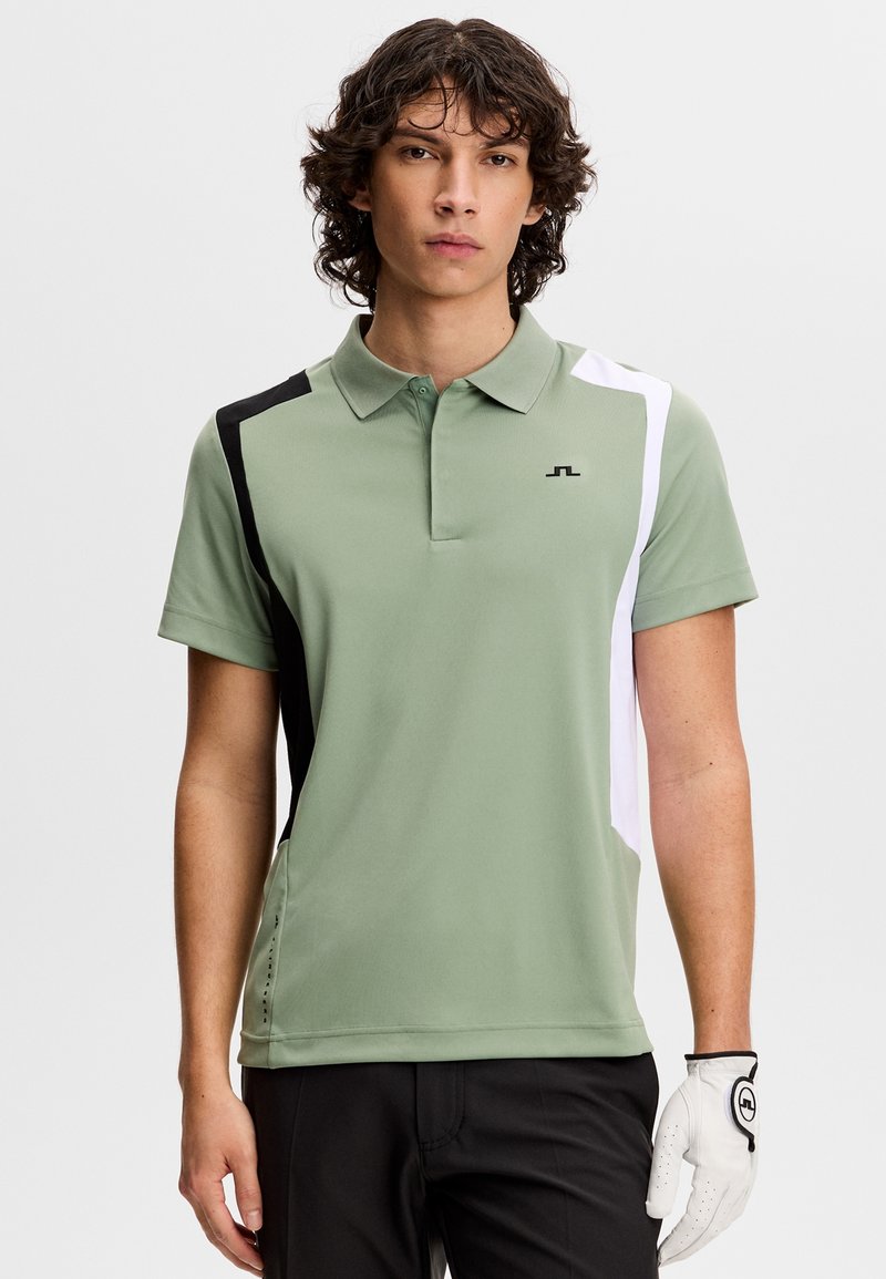 Jeune homme portant un polo vert clair avec des panneaux noirs et blancs, un pantalon noir, et un gant de golf blanc à la main droite.