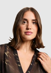 Blouse avec une base noire, présentant un léger motif abstrait. Fabriquée en tissu transparent, elle a un col en V et des manches amples. Le modèle touche le col.