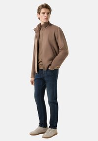 Brown fleece jacket with high collar over a beige top, paired with dark blue jeans and light beige shoes. Smooth texture, casual design.