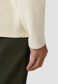 Off-white gebreide trui met geribbelde manchetten, weergegeven op een hand die rust nabij donkergroene stof, waarbij de textuur en kleurcontrasten worden benadrukt.