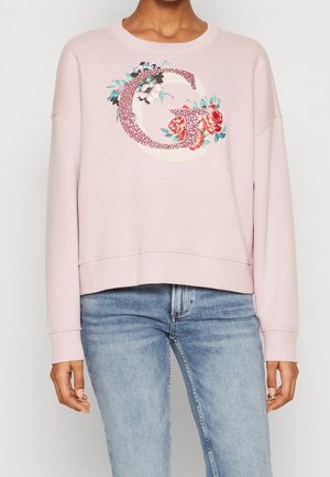 Mujer que lleva una sudadera rosa claro con una gran letra "G" floral en el frente, combinada con vaqueros azul claro, de pie frente a un fondo liso.