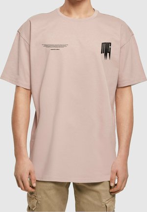 Mand iført en lyserød oversized t-shirt med lille sort tekst og en stiliseret sort "MC"-logo på brystet, kombineret med beige cargobukser.