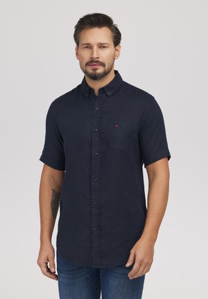 Mężczyzna o krótkich ciemnych włosach i brodzie, ubrany w granatową koszulę na guziki z krótkim rękawem oraz niebieskie dżinsy, stojący na jasnym tle.