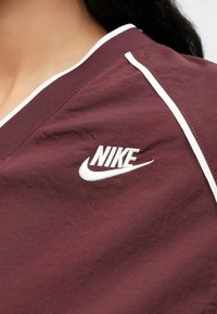 Vínový športový top s výstrihom do V. Má biely lem okolo goliera a po stranách. Na hrudi je vyšitý biely logotyp Nike.