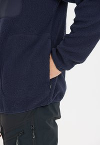 Marinblå fleece-pullover med en texturerad yta, som har en sidoficka och en mjuk tygfinish. Sömmar synliga längs nederkanten.