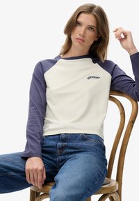 Młoda kobieta siedząca na drewnianym krześle, ubrana w białą i fioletową bluzkę z długim rękawem z logo "Superdry" oraz niebieskie dżinsy.