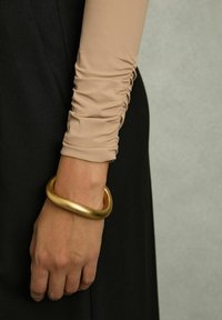 Gouden metalen armband om een hand, met een glad, rond ontwerp. De hand is gekleed in een lichtbeige top met ruches en lange mouwen.