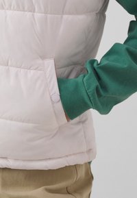 Helles rosa gepolstertes Weste mit sichtbarer Seitentasche, Knopfverschluss und grünem Langarmshirt darunter; weiche Textur und gestepptes Design.