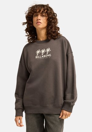 Frau mit lockigem Haar trägt einen dunklen, übergroßen Sweatshirt mit drei Palmen und "Billabong"-Logo auf weißem Hintergrund.