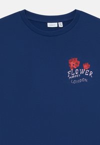 Ναυτικό μπλε πουλόβερ με στρογγυλή λαιμόκοψη, διακοσμημένο με κόκκινο λουλουδάτο σχέδιο και λευκό κείμενο "Flower Market London" στην επάνω αριστερή πλευρά του στήθους.