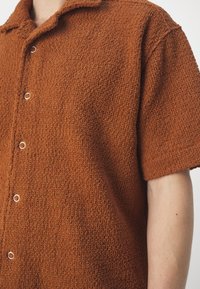 Gros plan d'une personne portant une chemise à manches courtes texturée de couleur rouille avec des boutons-pression argentés et un col.