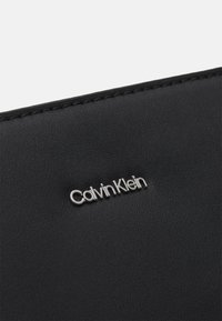 Svart plånbok i konstläder med präglad silverfärgad "Calvin Klein"-logotyp, med noggrant stickning längs kanterna och en slät yta.