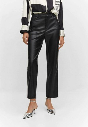 Femme portant un pantalon en cuir noir taille haute, un chemisier à motifs noir et blanc, et des talons pointus argentés avec des brides à la cheville.