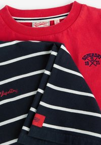 Rotes Baumwoll-T-Shirt mit geripptem Halsauschnitt, darunter ein navyblau gestreiftes Hemd. Beide tragen Logos in kontrastierenden Farben und die Etiketten sind sichtbar.
