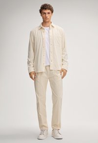 Beige doorgestikte overshirt met knoopsluiting, witte T-shirt eronder, gecombineerd met lichtbeige broek en witte sneakers.