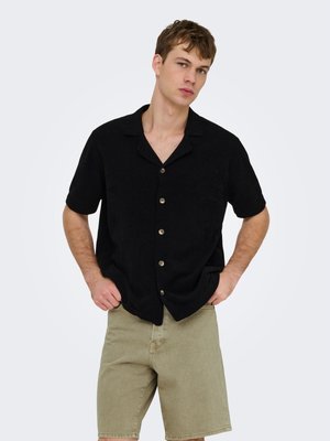 Ung mann som står med hendene i lommene, iført en kortermet svart knappeskjorte og beige knelang shorts på hvit bakgrunn.