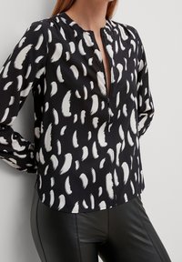 Blouse noire avec un motif de coups de pinceau abstrait blanc, décolleté en V, manches longues et un tissu léger. Associée à un pantalon noir.