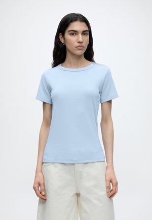 Kobieta o długich ciemnych włosach, ubrana w jasnoniebieską koszulkę z krótkim rękawem oraz szerokie kremowe spodnie, stoi na tle jednolitego białego tła.