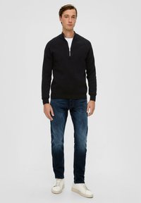 Schwarzer Reißverschluss-Pullover mit geripptem Strickmuster, kombiniert mit dunkelblauen Jeans und weißen Sneakers. Das Model steht aufrecht und blickt nach vorne.