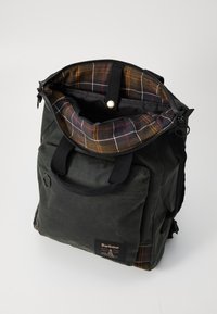 Dark green Barbour backpack with front pocket, black handles, zipper opened to show brown and orange plaid interior lining and snap closure strap.