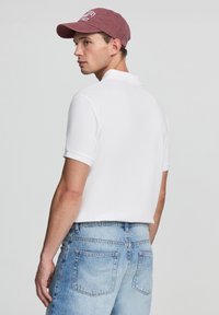 Biała krótka koszulka polo o prostym fasonie, zestawiona z jasnoniebieskimi szortami dżinsowymi. Całość dopełnia bordowa czapka z haftowanym napisem.