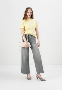 T-shirt jaune à manches courtes, jeans gris taille haute à jambes larges, et talons slingback beige à bout pointu, avec un petit sac à main blanc.