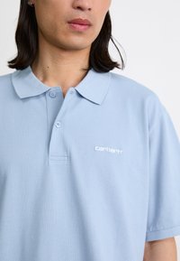 Polo bleu clair en mélange de coton, avec un col classique, une patte de boutonnage à trois boutons et un logo brodé discret sur la poitrine.