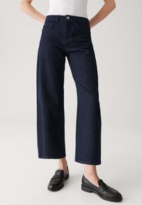 Donkerblauwe wijde spijkerbroeken van denim, met een klassiek vijf-pocketontwerp en een eenvoudige tailleband met een zilveren knoop. Zwarte instappers gedragen.