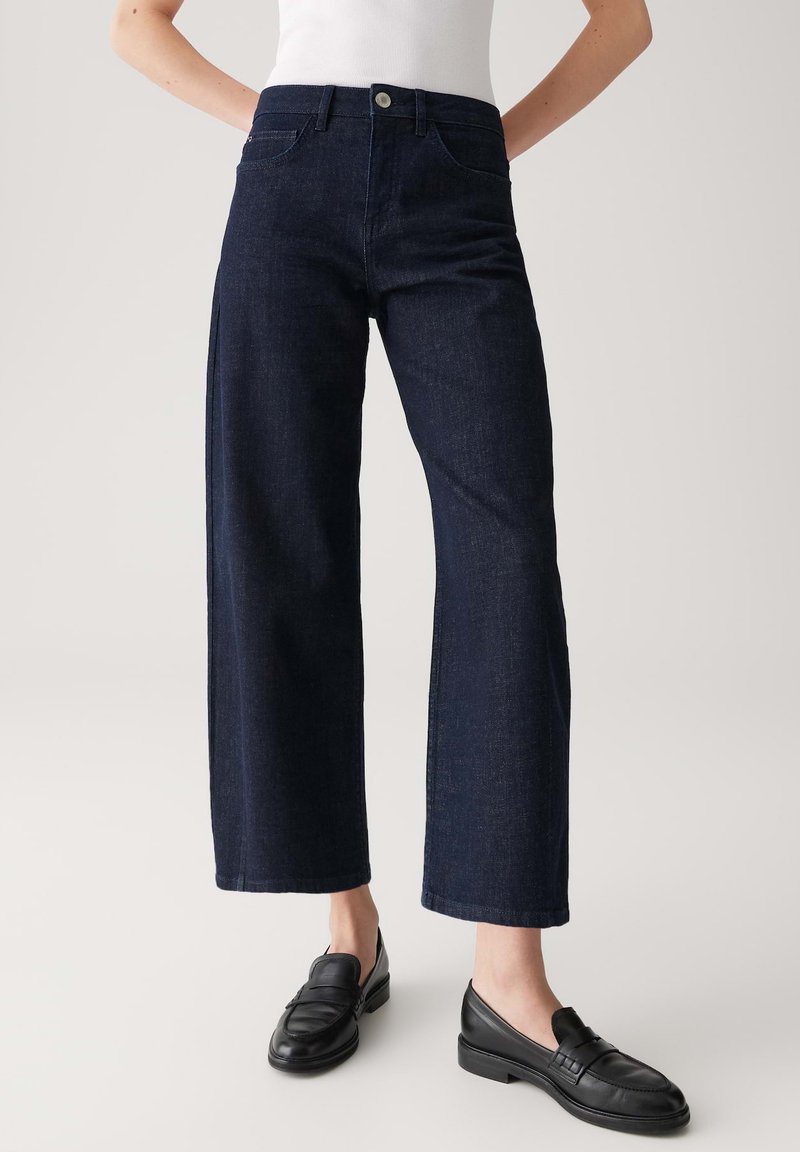 Donkerblauwe wijde spijkerbroeken van denim, met een klassiek vijf-pocketontwerp en een eenvoudige tailleband met een zilveren knoop. Zwarte instappers gedragen.