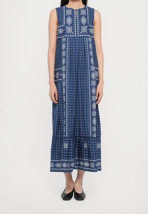 Mouwloze blauwe maxi-jurk met witte geometrische en bloemmotieven, gedragen met zwarte platte schoenen, tegen een effen achtergrond.