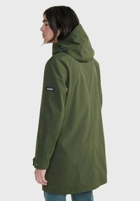 Cappotto impermeabile verde oliva con cappuccio, silhouette lunga e una patch del logo sulla manica sinistra. Texture liscia e opaca e polsini abbottonati.