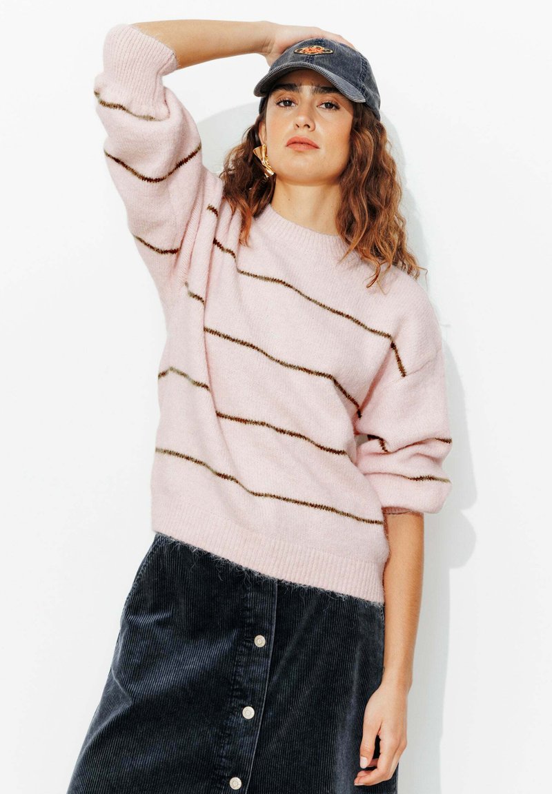 Femme portant un pull rayé rose, une jupe en velours côtelé foncé boutonnée et une casquette grise, posant avec un bras levé devant un fond blanc.